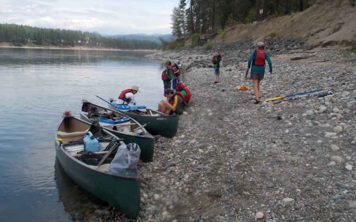 a few kayaks rest on the shore of a lake while people carry gear away from them 